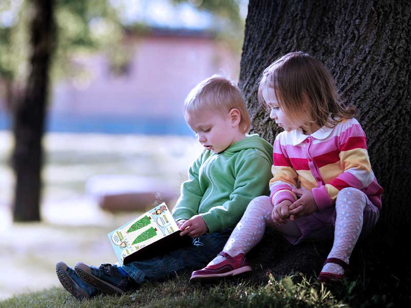 Two young children sitting under a tree outdoors, with one child holding and reading a Christmas trail activity booklet designed for English Heritage.