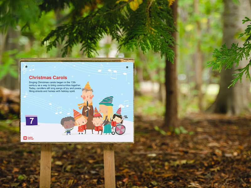 Christmas Carols trail panel installed on a wooden post in a woodland setting, featuring illustrated characters and educational content for an English Heritage seasonal trail.