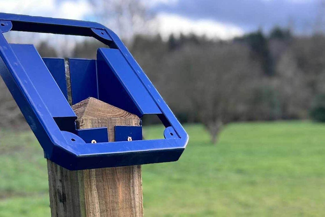 A close-up view of a blue metal trail marker frame fixed to a wooden post, showing the empty holder ready for interchangeable trail panels, with a grassy field and trees in the background.