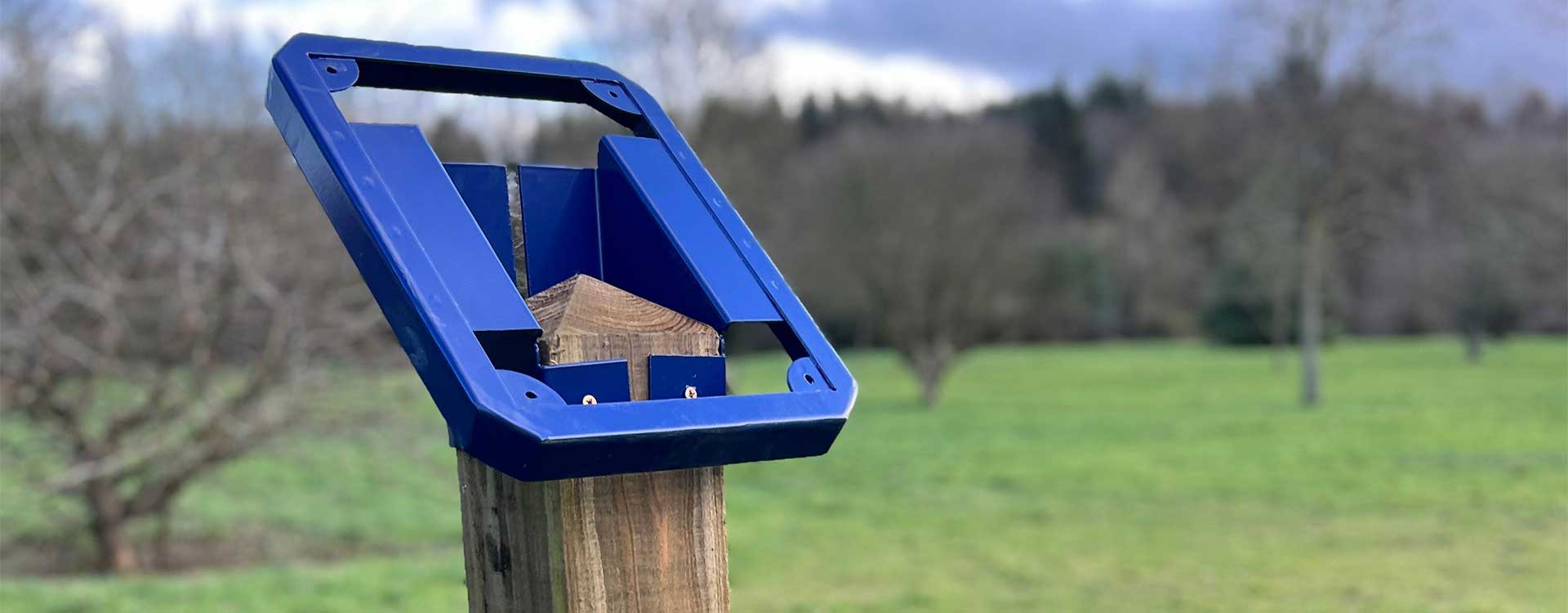A close-up view of a blue metal trail marker frame fixed to a wooden post, showing the empty holder ready for interchangeable trail panels, with a grassy field and trees in the background.